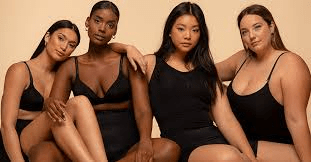 Diverse group of four women in black lingerie poses confidently against a neutral backdrop, promoting body positivity and inclusivity.