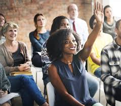 A diverse audience in a workshop, with a woman smiling and raising her hand to ask a question. The image highlights engagement in a learning environment.