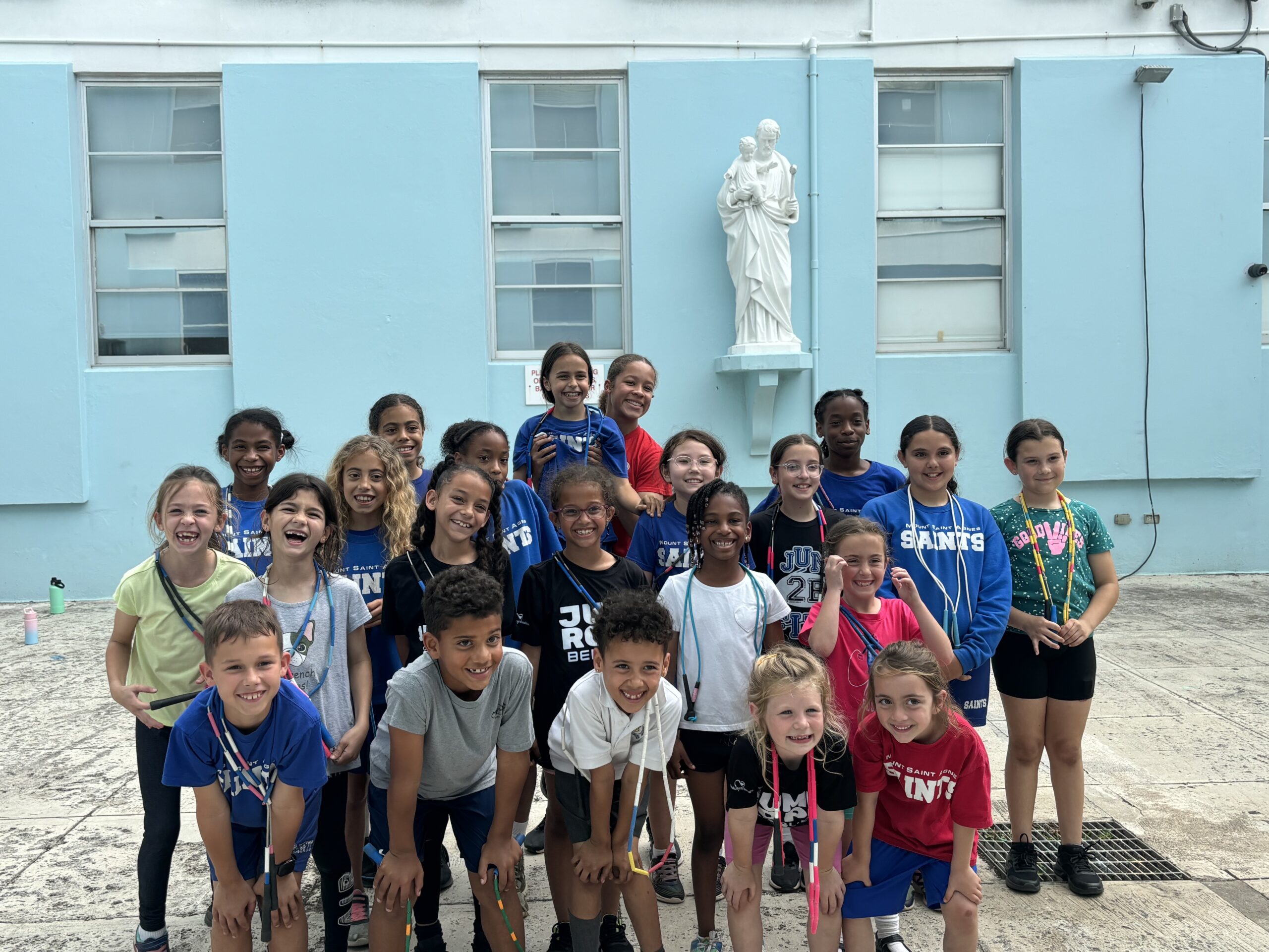 Group of smiling children gathered outdoors, posing cheerfully in front of a light blue wall and a statue, emphasizing teamwork and joy in their activities.