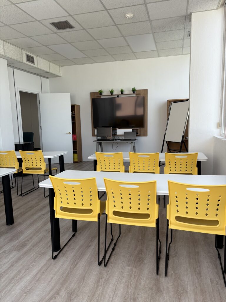 Classroom with neatly arranged white tables and yellow chairs facing a large screen, beneath three potted plants. Bright and organized atmosphere.