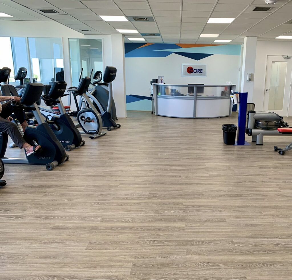Modern gym interior with wood flooring, exercise bikes on the left, and a sleek reception desk in front. The space is bright and inviting.
