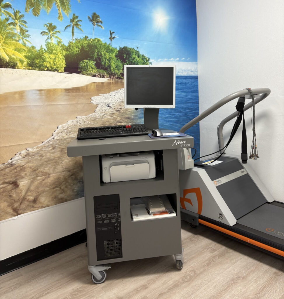 A treadmill next to a medical cart with a computer, set against a vibrant beach wall mural with palm trees and bright sunshine, creating a serene yet clinical atmosphere.